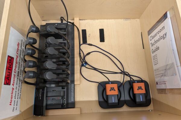 Pedestal - inside view of drawer showing two wireless mics in charging base and assistive listening devices in charger