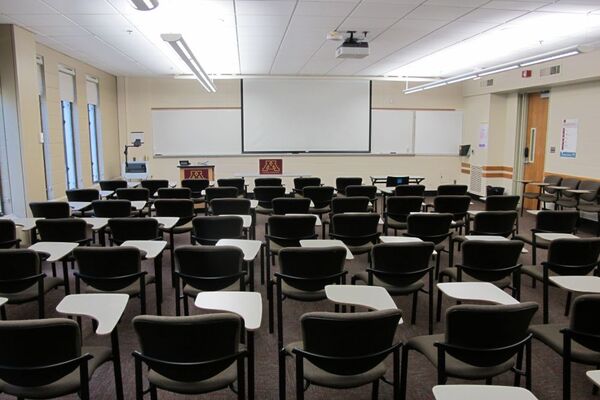 Front of room view with lectern on left in front of markerboard, projection screen lowered, and exit door on right wall