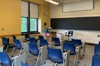 Front of room view with lectern on left in front of chalkboard and projection screen partially lowered