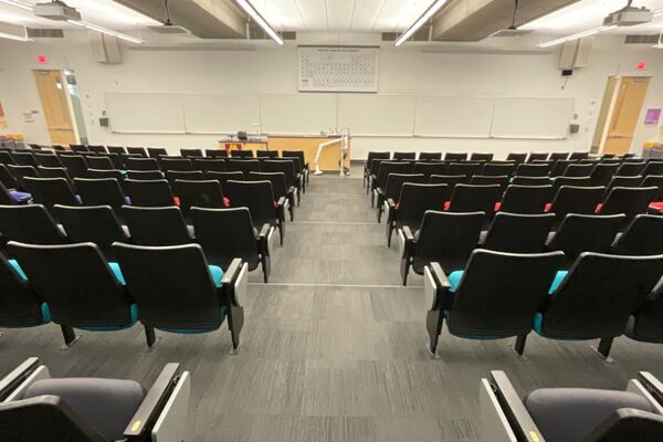 Front of room view with lectern on left of demonstration bench in front of markerboard, exit doors on left and right sides of front wall