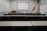 Front of room view with lectern on left in front of markerboard and projection screen lowered, exit door on right wall near front of room