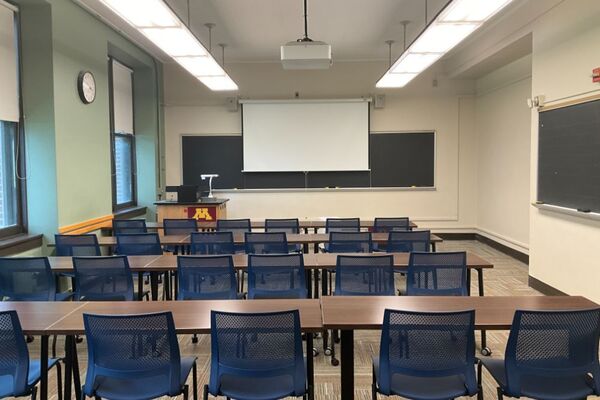 Front of room view with lectern on left in front of chalkboard and projection screen partially lowered