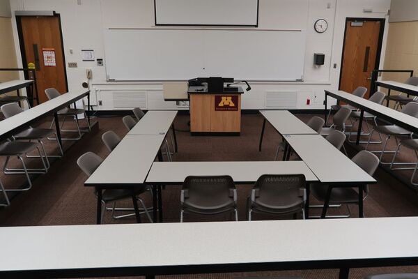 Front of room view with lectern center in front of markerboard and projection screen partially raised, exit doors on left and right of markerboard