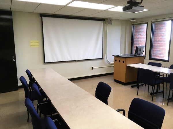 Photo of front of room from back of room, tables arranged in a U shape, windows on the right wall.