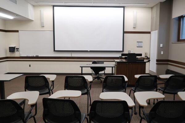 Front of room view with lectern on right in front of markerboard and projection screen partially lowered