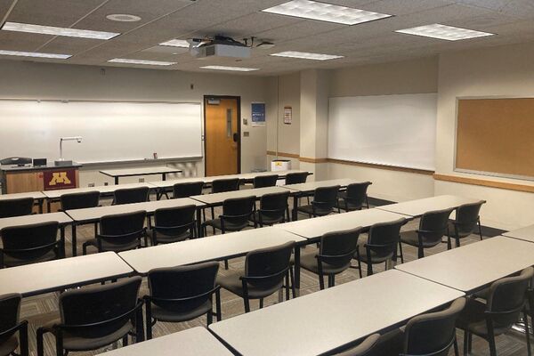 Front of room view with lectern on left in front of markerboard, exit door to the right, table and chair seating