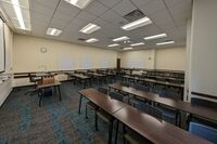 Back of room view of student table and chair seating, multiple markerboards on left side and back wall, exit door in left rear corner