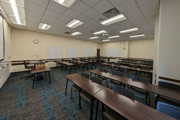 Back of room view of student table and chair seating, multiple markerboards on left side and back wall, exit door in left rear corner