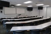 Front of room view with lectern on right in front of markerboard and display screens mounted on side walls