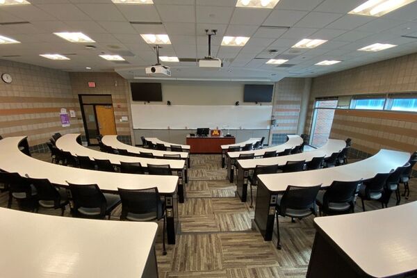 Front of room view with lectern center in front of markerboard and display screens to left and right, exit door to front left