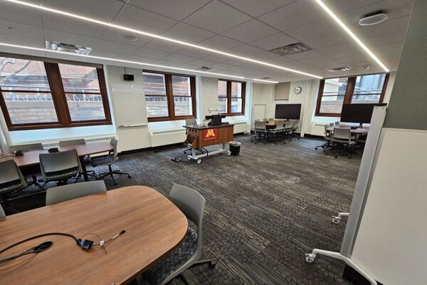 View of student active learning table and chair seating, multiple markerboards, and student display monitors at each table, instructor pedestal in center