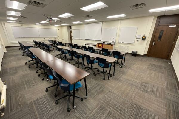 Front of room view with lectern on right in front of markerboards, additional markerboards on left side wall of room, and doors located in both corners of room