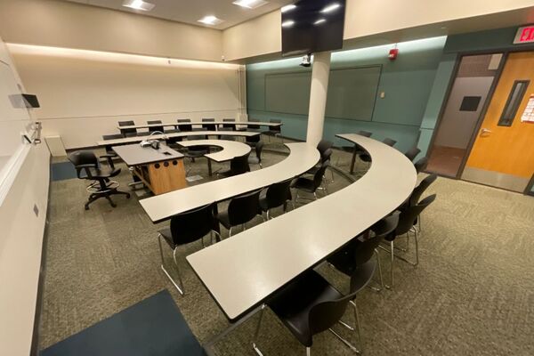 Back of room view of student tiered fixed-table and chair seating and confidence monitor on wall above