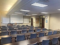 Front of room view with lectern on left in front of markerboard and projection screen lowered, exit door on the right