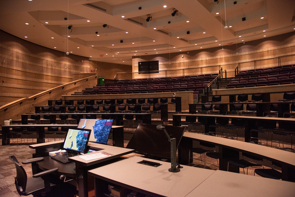 Instructor view from front right of classroom showing podium monitor, classroom camera centered in the room, two confidence panels at the front middle of the room, a document camera, and audience seating.  Ceiling mounted audience mics are also visible.