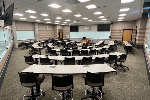 Front of room view with lectern center in front of markerboard, exit door on front left, chairs and fixed tables 