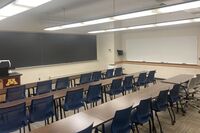 Front of room view with lectern on left in front of chalkboard, projection screen lowered