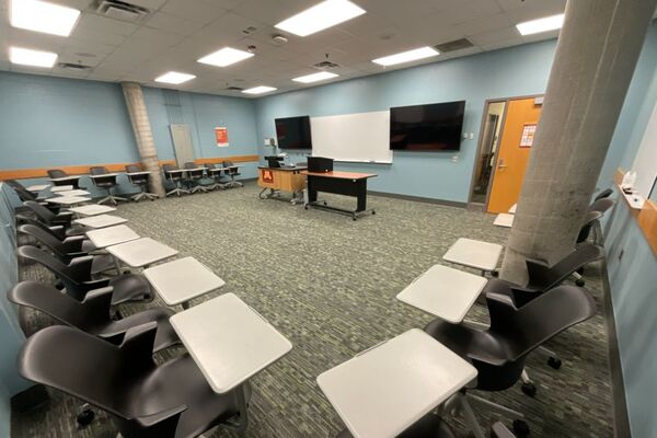 Front of room view with lectern center in front of markerboard and display screens to the left and right, exit door on the right