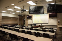 Audience view from right rear. Two displays next to front projector display behind the podium are visible, and the rear of the confidence panels can be seen.  Audience seating is visible.  Exit door is on the far left side.