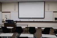 Front of room view with lectern on left in front of markerboard and projection screen partially lowered