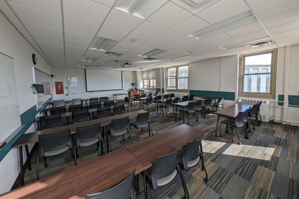 Front of room view with lectern on right in front of markerboard and projection screen lowered, exit door in left corner of room