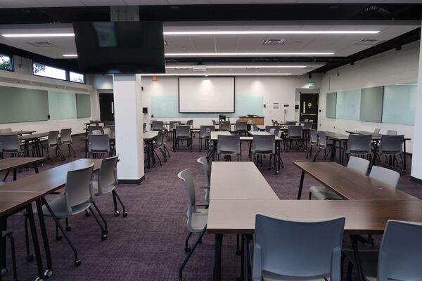 Front of room view with lectern on right in front of markerboard, exit doors on left and right, and projection screen lowered