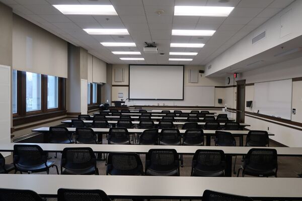 Front of room view with lectern on left in front of markerboard and projection screen lowered, exit door on front right wall