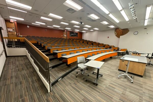 Back of room view of student tiered fixed table and chair seating, exit doors on right and left, and A/V booth on right