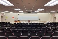 Front of room view with lectern on left in front of markerboard and exit doors on left and right